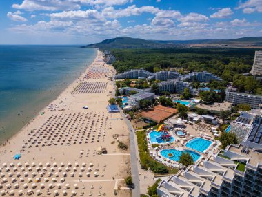 Sırayla dizilmiş şemsiyeler, güneş banyosu ve su aktiviteleriyle dolu geniş bir plajın insansız hava aracı videosu. Hava manzarası, deniz kıyısındaki tatil beldesindeki bir yaz gününün canlı atmosferini yakalar. Albena, Bulgaristan.