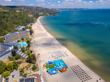 Sırayla dizilmiş şemsiyeler, güneş banyosu ve su aktiviteleriyle dolu geniş bir plajın insansız hava aracı videosu. Hava manzarası, deniz kıyısındaki tatil beldesindeki bir yaz gününün canlı atmosferini yakalar. Albena, Bulgaristan.