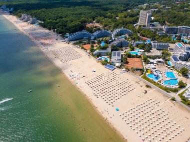 Sırayla dizilmiş şemsiyeler, güneş banyosu ve su aktiviteleriyle dolu geniş bir plajın insansız hava aracı videosu. Hava manzarası, deniz kıyısındaki tatil beldesindeki bir yaz gününün canlı atmosferini yakalar. Albena, Bulgaristan.