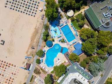 Sırayla dizilmiş şemsiyeler, güneş banyosu ve su aktiviteleriyle dolu geniş bir plajın insansız hava aracı videosu. Hava manzarası, deniz kıyısındaki tatil beldesindeki bir yaz gününün canlı atmosferini yakalar. Albena, Bulgaristan.