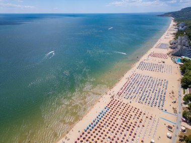 Sırayla dizilmiş şemsiyeler, güneş banyosu ve su aktiviteleriyle dolu geniş bir plajın insansız hava aracı videosu. Hava manzarası, deniz kıyısındaki tatil beldesindeki bir yaz gününün canlı atmosferini yakalar. Albena, Bulgaristan.