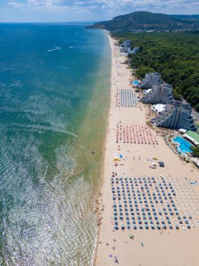 Sırayla dizilmiş şemsiyeler, güneş banyosu ve su aktiviteleriyle dolu geniş bir plajın insansız hava aracı videosu. Hava manzarası, deniz kıyısındaki tatil beldesindeki bir yaz gününün canlı atmosferini yakalar. Albena, Bulgaristan.