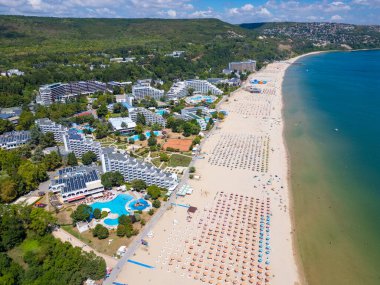 Sırayla dizilmiş şemsiyeler, güneş banyosu ve su aktiviteleriyle dolu geniş bir plajın insansız hava aracı videosu. Hava manzarası, deniz kıyısındaki tatil beldesindeki bir yaz gününün canlı atmosferini yakalar. Albena, Bulgaristan.