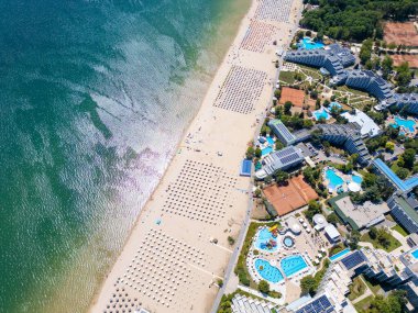 Sırayla dizilmiş şemsiyeler, güneş banyosu ve su aktiviteleriyle dolu geniş bir plajın insansız hava aracı videosu. Hava manzarası, deniz kıyısındaki tatil beldesindeki bir yaz gününün canlı atmosferini yakalar. Albena, Bulgaristan.