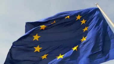 Avrupa Birliği bayrağının ikonik mavi arkaplanı ve altın yıldızlarıyla rüzgarda dalgalanan yakın plan çekimi. Kumaşın hareketi dinamik ve vatansever bir hava katıyor..