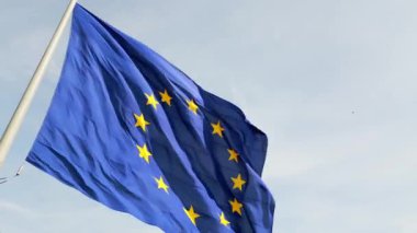 Avrupa Birliği bayrağının ikonik mavi arkaplanı ve altın yıldızlarıyla rüzgarda dalgalanan yakın plan çekimi. Kumaşın hareketi dinamik ve vatansever bir hava katıyor..