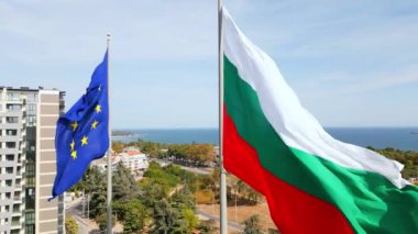 Avrupa Birliği ve Bulgar bayrakları kıyı kenti üzerinde yan yana dalgalanırken, arka planda deniz ve kentsel manzara bulunuyor. Görüntü birlik ve milli gururu simgeliyor..