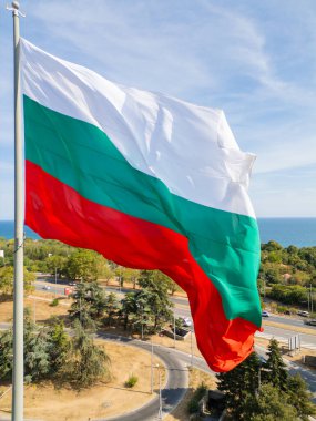 Bulgar bayrağı, arka planında engin deniz ve yeşil manzarayla bir kıyı kenti üzerinde gururla dalgalanıyor. Ulusal gurur ve güzel doğal manzaranın sembolü..