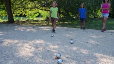 Bir aile güneşli bir parkta petanque oynuyor. Anne, iki oğlunun metal topları hedefe doğru atmasını izliyor. Birlikte hava hareketlerinin keyfini çıkarıyorlar. Toplara odaklan.