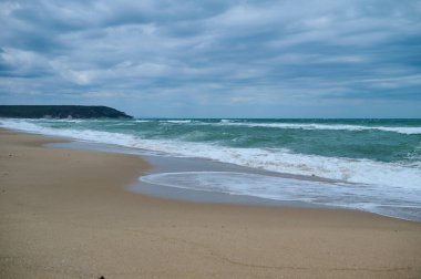 Dalgalar sakin, kumlu bir sahile çakılırken dramatik bir gökyüzü tepemizde beliriyor. Rahatlama ve tefekkür için mükemmel huzurlu bir atmosfer yaratıyor..