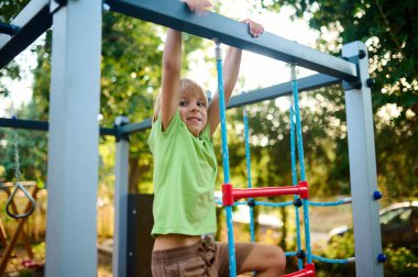 Genç bir çocuk, güneşli bir parkta oynamaktan zevk alırken gülümseyen bir tırmanış çerçevesinden sarkıyor. Ağaçlar doğal bir zemin sağlar, neşeli atmosferi güçlendirir..
