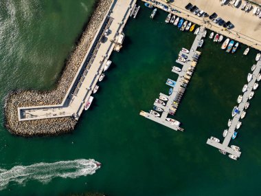 Bir marinaya çok sayıda tekne yanaşmış ve bazıları uzun bir rıhtım boyunca demirlemiş. Su sakin, ve güneş gölgeleri salıyor, canlı sahneyi güçlendiriyor..