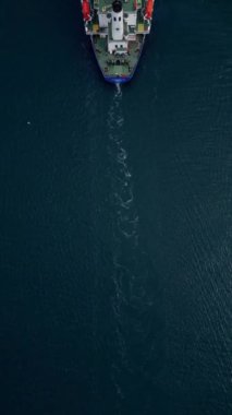 Renkli konteynırlarla dolu büyük bir konteynır gemisi geniş bir derin mavi su boyunca yelken açar. Gemi arkasında küçük bir iz bırakır.