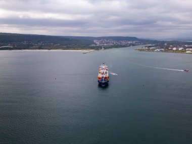 A large cargo container ship moves through the harbor waters, flanked by a smaller boat heading toward the shoreline. The backdrop features a coastal landscape under overcast skies. clipart