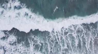 Su dinamik bir şekilde çalkalanır dalgalar kumlu sahile doğru yuvarlanır, beyaz köpük üretir ve güneşin altında okyanusun canlı mavi renklerini gözler önüne serer..