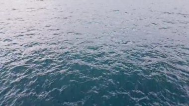Sakin bir okyanus yüzeyi, berrak gökyüzünün altındaki güneş ışığını yansıtır ve suyun sakinliğini vurgular. Sahne öğle vakti huzur ve dinginlik hissi veriyor..