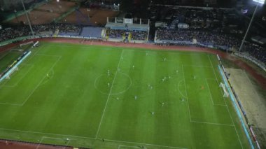 İnsansız hava aracı, gece devam eden bir futbol maçının havadan görüntüsünü yakalıyor. Oyuncuları parlak bir sahada ve kalabalık bir stadyumda gösteriyor..