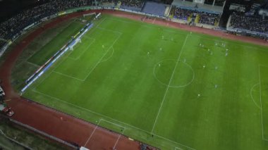 İnsansız hava aracı, gece devam eden bir futbol maçının havadan görüntüsünü yakalıyor. Oyuncuları parlak bir sahada ve kalabalık bir stadyumda gösteriyor..