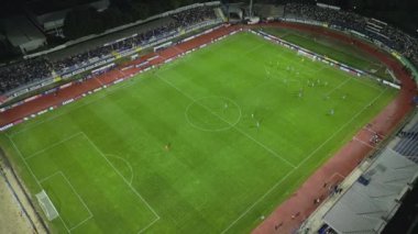 İnsansız hava aracı, gece devam eden bir futbol maçının havadan görüntüsünü yakalıyor. Oyuncuları parlak bir sahada ve kalabalık bir stadyumda gösteriyor..