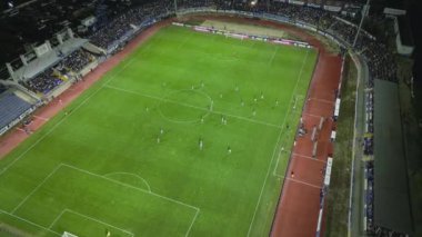 İnsansız hava aracı, gece devam eden bir futbol maçının havadan görüntüsünü yakalıyor. Oyuncuları parlak bir sahada ve kalabalık bir stadyumda gösteriyor..