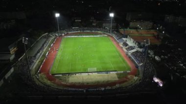 İnsansız hava aracı, parlak aydınlatılmış bir stadyumda gece maçını çekiyor. Seyirciler tribünleri doldururken takımlar sahada yarışıyor ve şehirde elektrik atmosferi yaratıyorlar..