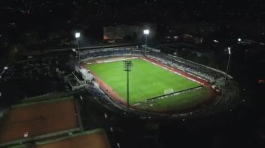 İnsansız hava aracı, parlak aydınlatılmış bir stadyumda gece maçını çekiyor. Seyirciler tribünleri doldururken takımlar sahada yarışıyor ve şehirde elektrik atmosferi yaratıyorlar..