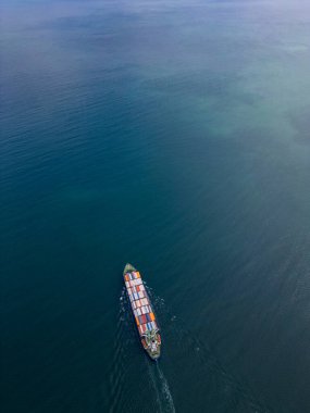 Renkli konteynırlarla dolu büyük bir konteynır gemisi geniş bir derin mavi su boyunca yelken açar. Gemi arkasında küçük bir iz bırakır.