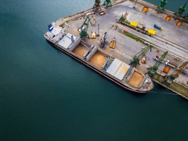 Limana yanaşan kargo gemisi vinçler açık ambardan tahıl kaldırırken yükleniyor. Çevreleyen makineler ve ekipmanlar görünür.