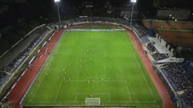 İnsansız hava aracı, parlak aydınlatılmış bir stadyumda gece maçını çekiyor. Seyirciler tribünleri doldururken takımlar sahada yarışıyor ve şehirde elektrik atmosferi yaratıyorlar..