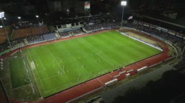 İnsansız hava aracı, parlak aydınlatılmış bir stadyumda gece maçını çekiyor. Seyirciler tribünleri doldururken takımlar sahada yarışıyor ve şehirde elektrik atmosferi yaratıyorlar..