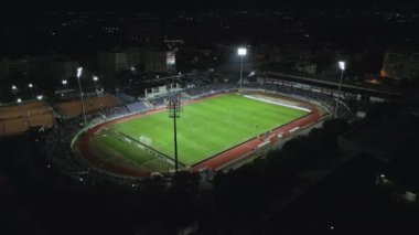 İnsansız hava aracı, parlak aydınlatılmış bir stadyumda gece maçını çekiyor. Seyirciler tribünleri doldururken takımlar sahada yarışıyor ve şehirde elektrik atmosferi yaratıyorlar..