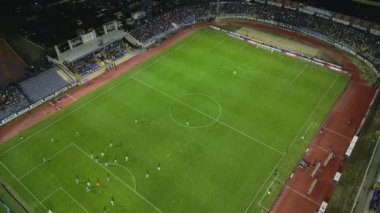 İnsansız hava aracı, parlak aydınlatılmış bir stadyumda gece maçını çekiyor. Seyirciler tribünleri doldururken takımlar sahada yarışıyor ve şehirde elektrik atmosferi yaratıyorlar..