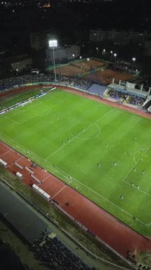 İnsansız hava aracı, parlak aydınlatılmış bir stadyumda gece maçını çekiyor. Seyirciler tribünleri doldururken takımlar sahada yarışıyor ve şehirde elektrik atmosferi yaratıyorlar..