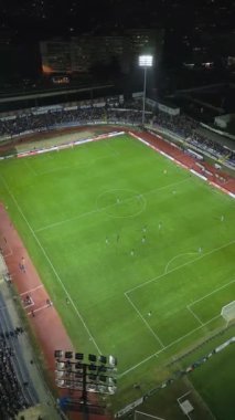 İnsansız hava aracı, parlak aydınlatılmış bir stadyumda gece maçını çekiyor. Seyirciler tribünleri doldururken takımlar sahada yarışıyor ve şehirde elektrik atmosferi yaratıyorlar..