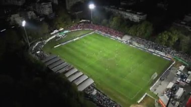 İnsansız hava aracı, parlak aydınlatılmış bir stadyumda gece maçını çekiyor. Seyirciler tribünleri doldururken takımlar sahada yarışıyor ve şehirde elektrik atmosferi yaratıyorlar..