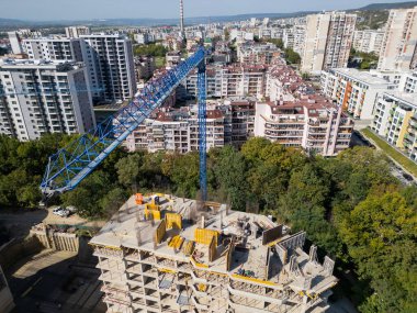 Bir vinç ve yakınlardaki konut binaları olan bir inşaat alanının insansız hava aracı videosu. Görüntüler yeşil ve şehir hayatı ile çevrili kentsel gelişimin ilerlemesini yakalıyor..