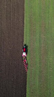 Kırmızı bir traktörün tarlayı sürüşünün havadan görüntüsü yeşil çimen ve kara toprak arasında açık bir çizgi oluşturuyor. Tarım makineleri tarım arazisinde faaliyet gösteriyor.
