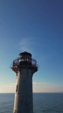 Kayalık dalgakıran üzerinde bir deniz fenerinin insansız hava aracı görüntüleri, mavi gökyüzü, sakin su ve yeşil yamaçlı deniz manzarası, sakin ve güneşli kıyı manzarası..