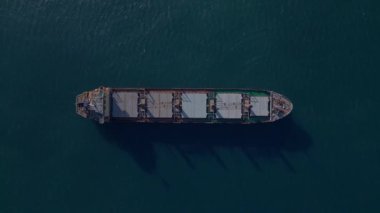 Açık denizde giden büyük bir kargo gemisinin tepeden aşağı hava aracı görüntüsü deniz yüzeyinde endüstriyel detayları ve gölgeleri gösteriyor. Denizcilik, nakliye ve lojistik temalar için ideal..
