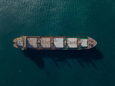 Açık denizde giden büyük bir kargo gemisinin tepeden aşağı hava aracı görüntüsü deniz yüzeyinde endüstriyel detayları ve gölgeleri gösteriyor. Denizcilik, nakliye ve lojistik temalar için ideal..