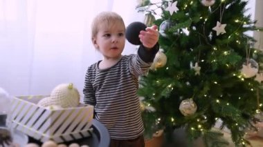Genç bir çocuk güzel aydınlatılmış bir Noel ağacını ışıldayan süslemelerle süslüyor. Yeni yürümeye başlayan çocuk meşgul ve meraklı. İçerideki bayram ruhunun tadını çıkarıyor..