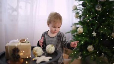 Genç bir çocuk güzel aydınlatılmış bir Noel ağacını ışıldayan süslemelerle süslüyor. Yeni yürümeye başlayan çocuk meşgul ve meraklı. İçerideki bayram ruhunun tadını çıkarıyor..