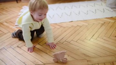 Neşeli genç bir çocuk yerde tahta bir oyuncakla oynuyor, sürünüyor ve keşfediyor. Bebek meşgul, meraklı ve sıcak bir yuva ortamında eğlenceli bir anın tadını çıkarıyor..