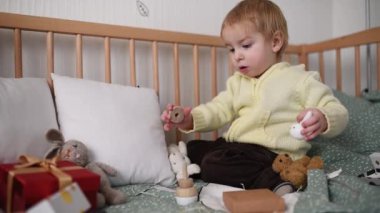 Küçük bir çocuk yatakta oturmuş, tahta bir oyuncağı merakla ve dikkatle tavşan şeklinde birleştiriyor. Etrafı yumuşak oyuncaklar ve hediye kutusuyla çevrili çocuk, huzurlu bir oyun zamanının keyfini çıkarıyor.