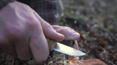 Bir adam ateşi bir bıçak ve yangın başlatıcı kullanarak tutuşturur, odun talaşlarının üzerinde kıvılcımlar yaratır. Bu yakın çekim hayatta kalma becerilerinin ve açık hava çalılığının özünü yakalar..