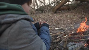 Genç bir çocuk ormanda kamp ateşinin yanında oturuyor, bıçakla odun oyuyor. Bu video doğada hayatta kalma becerileri ve çalı çırpı öğrenme ruhunu yakalıyor..
