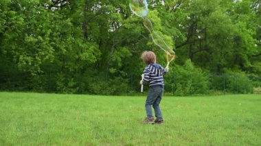 Çizgili tişörtlü mutlu bir çocuk yeşil bir parkta baloncuklu değnekle büyük renkli baloncuklar yaratıyor. O an, çocukluğun neşesini, neşesini ve güzelliğini yakalar..