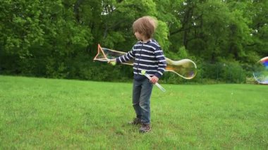 Çizgili tişörtlü mutlu bir çocuk yeşil bir parkta baloncuklu değnekle büyük renkli baloncuklar yaratıyor. O an, çocukluğun neşesini, neşesini ve güzelliğini yakalar..