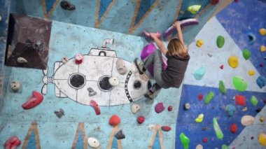 10 yaşında bir çocuk renkli tutacağı olan bir kaya duvarına tırmanıyor. Meydan okumaya odaklanmış, ustalıkla yukarı doğru hareket ediyor..