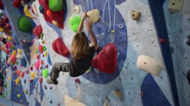 10 yaşında bir çocuk renkli tutacağı olan bir kaya duvarına tırmanıyor. Meydan okumaya odaklanmış, ustalıkla yukarı doğru hareket ediyor..